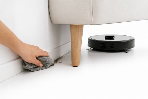 Una mano humana utiliza tranquilamente un pequeño paño para limpiar una esquina estrecha entre el rodapié y la pata de un mueble donde se ha acumulado algo de suciedad. Bastante más atrás permanece detenido un robot doméstico de limpieza que físicamente no puede acceder a ese pequeño rincón. El foco está en la mano y en la zona difícil, no en el robot. La escena transmite una limitación cotidiana de forma sencilla y honesta