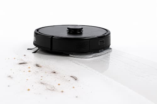 Delante del robot hay unas pocas migas, polvo fino y varios pelos de mascota claramente visibles; detrás de él, el suelo aparece perfectamente limpio. Cerca, unas pequeñas huellas húmedas muy sutiles muestran que también está realizando una pasada ligera de mopa. La escena comunica visualmente mantenimiento diario y no una limpieza profunda extrema.