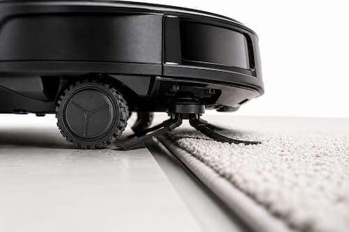 El detalle que realmente genera ruido: la rueda flexible y el pequeño cepillo lateral de un robot doméstico de limpieza atravesando suavemente una junta entre suelo duro y una alfombra fina. La imagen muestra con enorme detalle los materiales blandos, las pequeñas ruedas amortiguadas y el contacto delicado con el suelo. El robot completo no aparece; solamente los elementos mecánicos relacionados con vibraciones y contacto