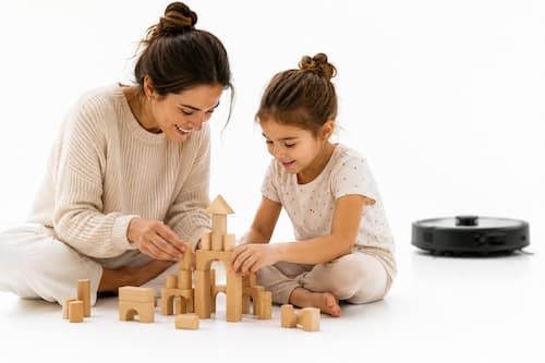 Una mujer adulta está sentada cómodamente en el suelo junto a una niña construyendo juntas una estructura con piezas de madera, completamente concentradas y disfrutando del momento. Muy al fondo y ligeramente desenfocado, un pequeño robot doméstico realiza discretamente una tarea de limpieza. La tecnología ocupa menos del diez por ciento de la imagen y nunca compite visualmente con las personas. El protagonismo absoluto pertenece al tiempo compartido y a la tranquilidad.