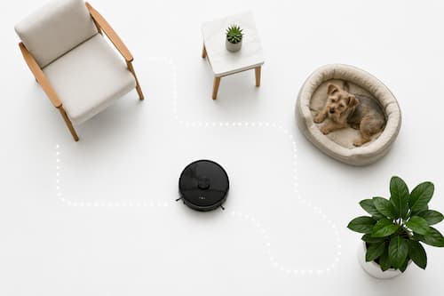 Un robot doméstico pequeño recorre un espacio minimalista organizado mediante pocos elementos reales: una silla, una pequeña mesa, una cama para perro y una planta. La distribución permite intuir visualmente un recorrido limpio y ordenado alrededor de los objetos